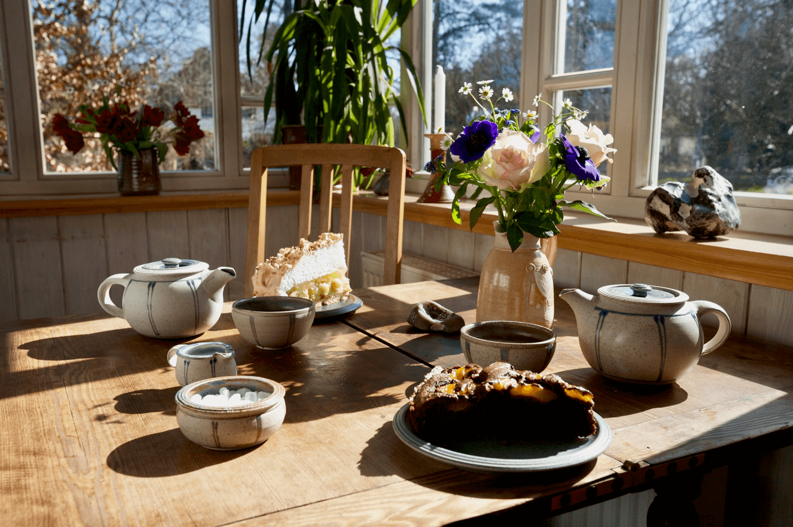 Sonnendurchfluteter Holztisch in der Teestube der Teeschale Prerow mit Teekannen, Tassen, zwei Kuchentellern und einer Blumenvase – gemütliche Café-Atmosphäre mit Ostsee-Charme.