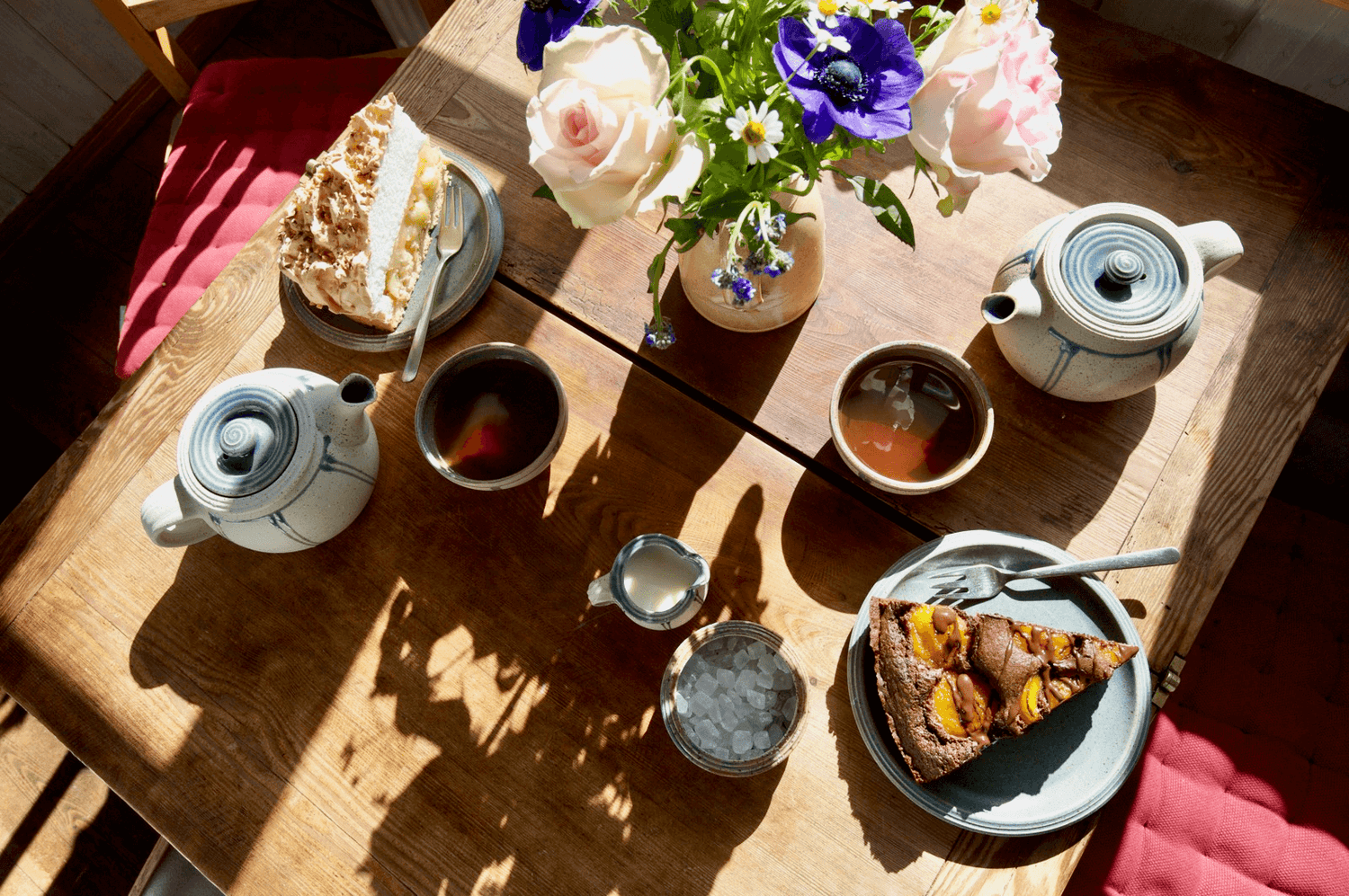 Gedeckter Holztisch in der Teestube der Teeschale Prerow mit Teekannen, Tassen, Kuchen und frischen Blumen im Sonnenlicht – gemütliche Café-Atmosphäre mit Ostsee-Charme.